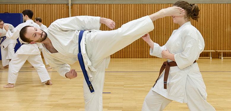 Zwei Karateka in traditioneller Kleidung üben Kicks und Techniken in einer Sporthalle während eines Trainings.