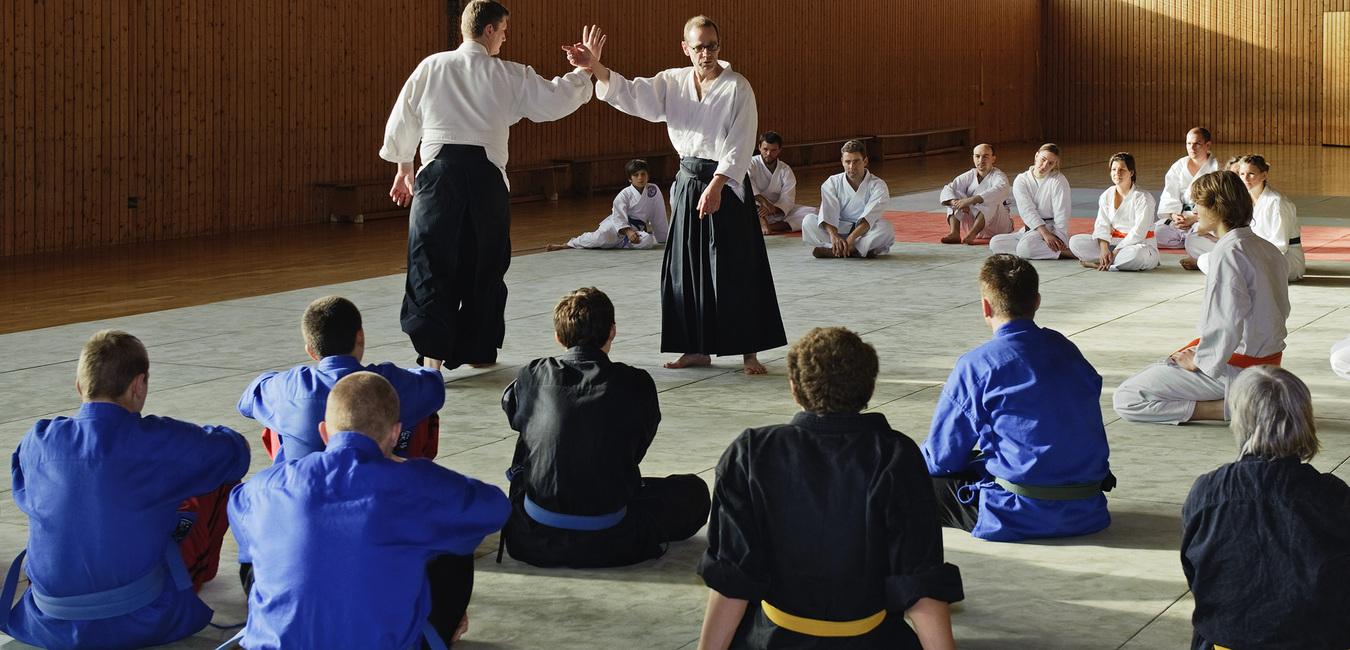 Zwei Judolehrer demonstrieren Techniken vor einer Gruppe von Schülern auf der Matte in einem Trainingsraum.