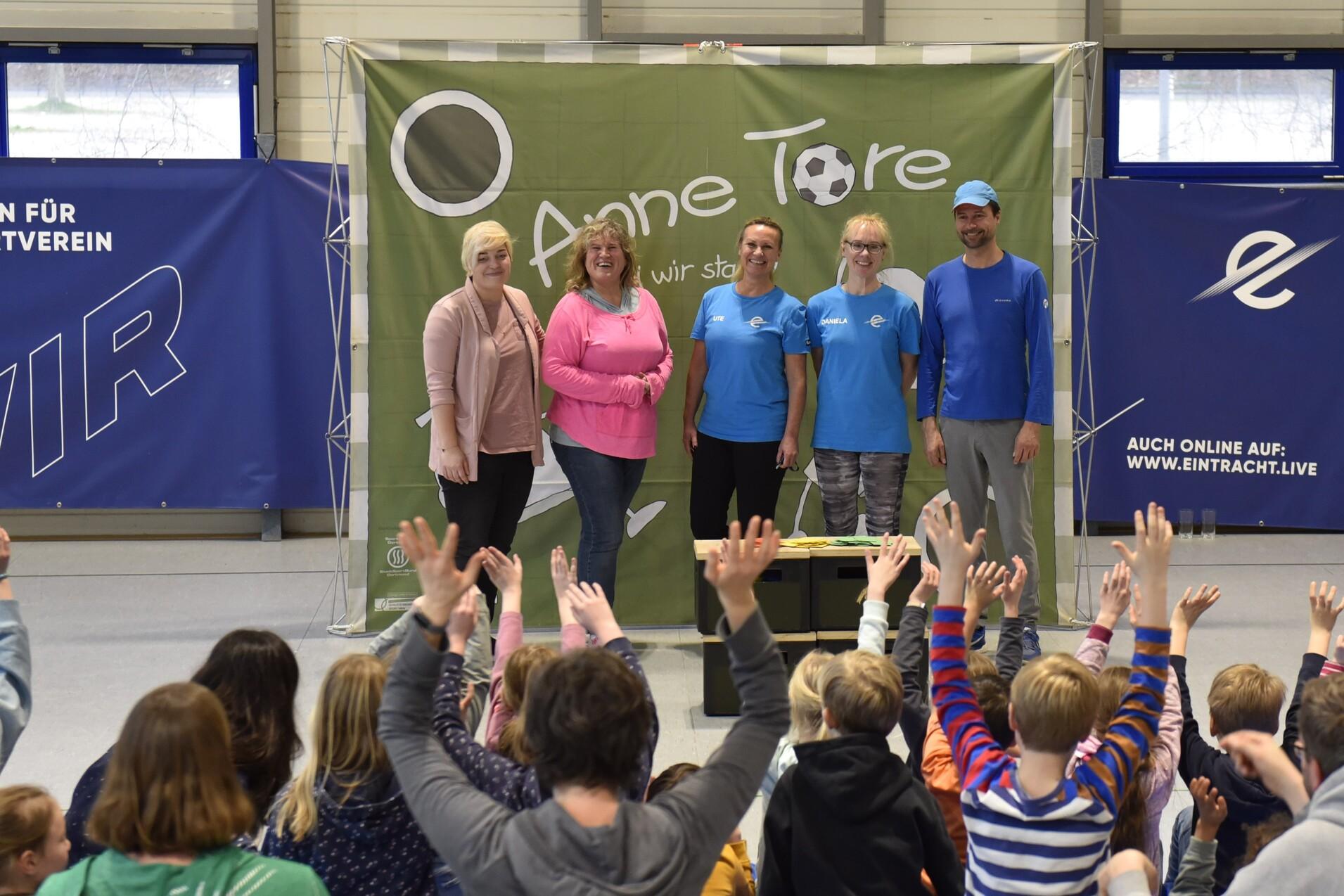 Gruppierung von vier Personen vor einem Banner, w&auml;hrend Kinder im Hintergrund die H&auml;nde heben. Sporthallenatmosph&auml;re.