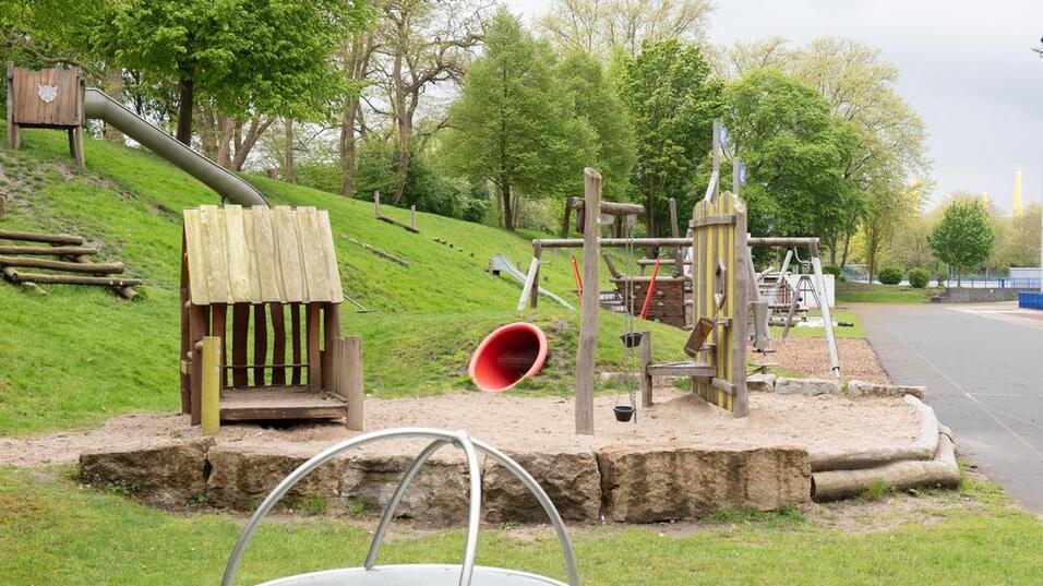 Spielplatz mit Holzspielger&auml;ten, Rutsche und Sandbereich, umgeben von B&auml;umen und Rasenfl&auml;chen.