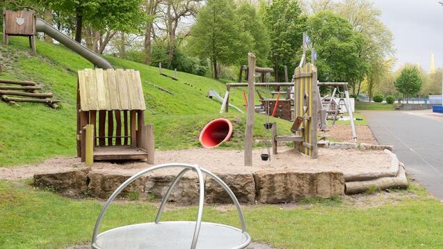 Spielplatz mit Holzspielgeräten, Rutsche und Sandbereich, umgeben von Bäumen und Rasenflächen.