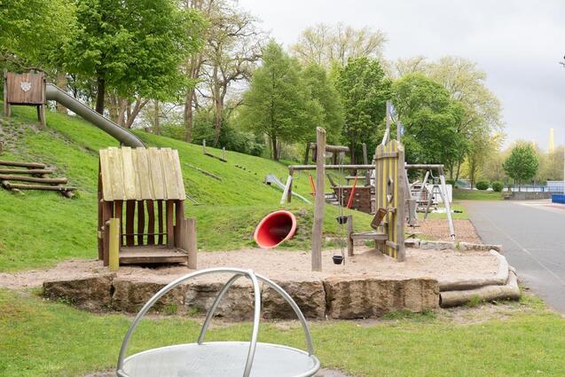 Spielplatz mit Holzspielgeräten, Rutsche und Sandbereich, umgeben von Bäumen und Rasenflächen.