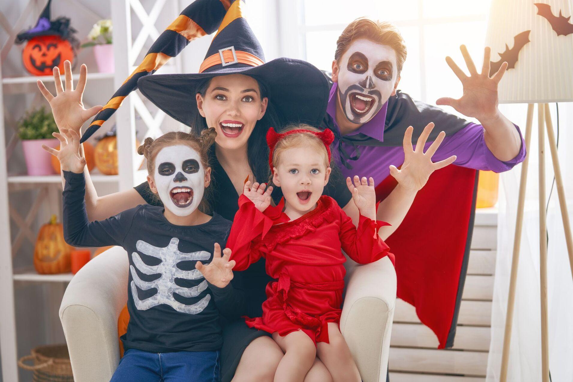 Familie in Halloween-Kostümen sitzt im Wohnzimmer, überzeugt lachend mit gruseligen Gesichtsmasken und Gesten.