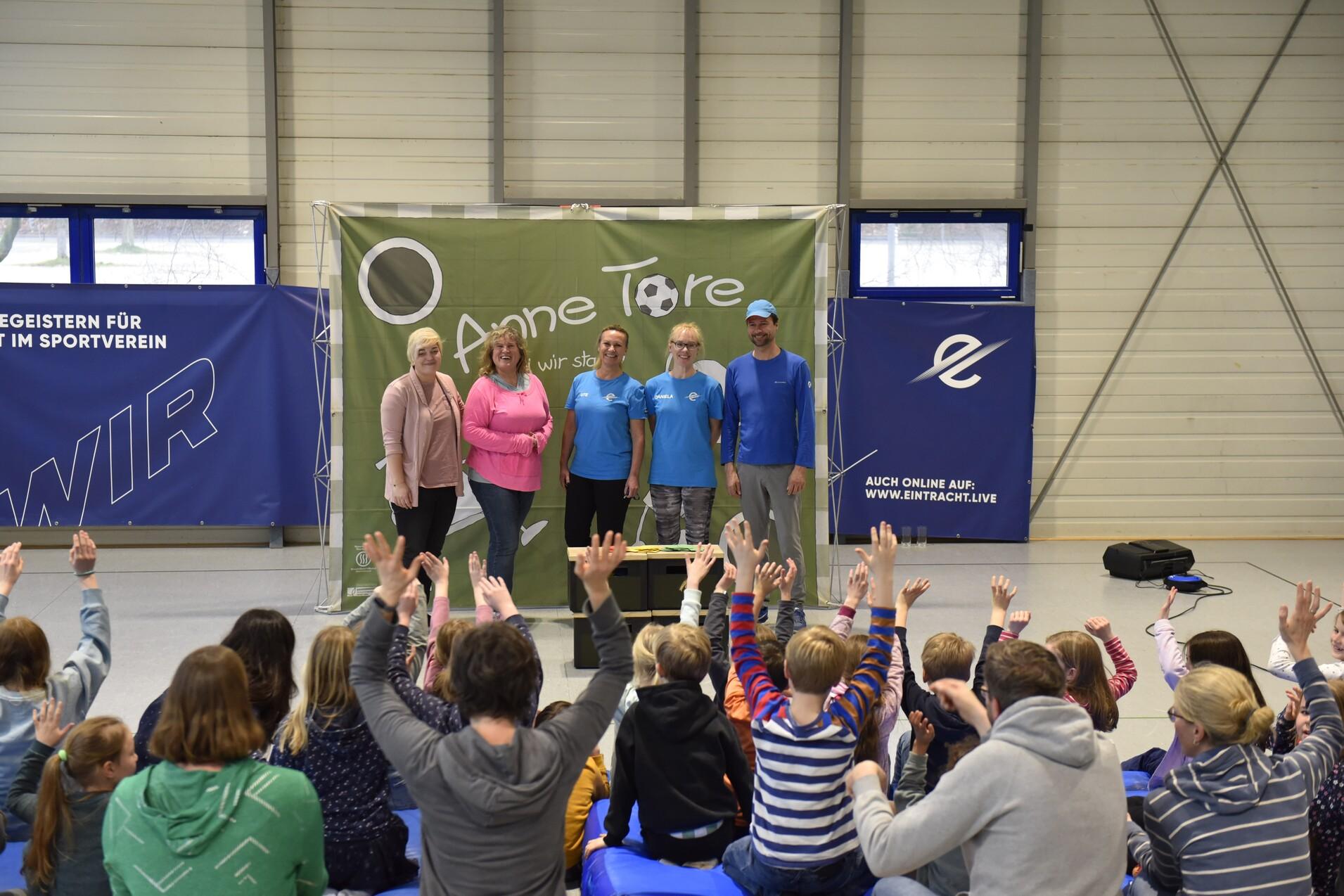 Gruppierung von vier Personen vor einem Banner, während Kinder im Hintergrund die Hände heben. Sporthallenatmosphäre.