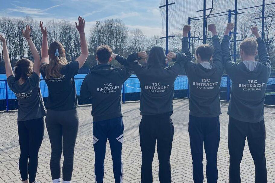 Gruppe von fünf Personen in Sweatshirts, die mit erhobenen Händen auf einem Sportgelände stehen. Text: „FSJler*innen gesucht!“