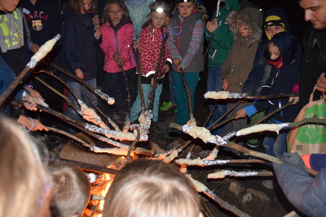 Gruppierung von Kindern um ein Lagerfeuer, w&auml;hrend sie Stockbrot an St&ouml;cken r&ouml;sten. Nachtaufnahme mit flammendem Feuer.