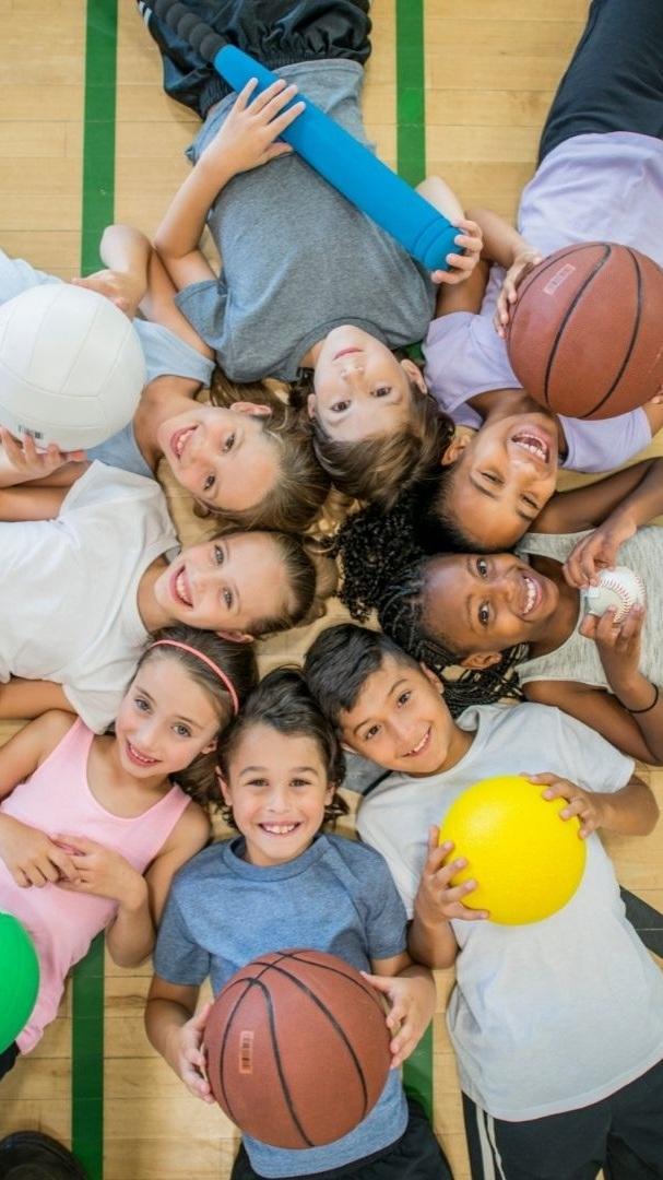 Gruppe von acht Kindern liegend auf einem Sportboden, umgeben von verschiedenen Sportb&auml;llen und -ger&auml;ten.
