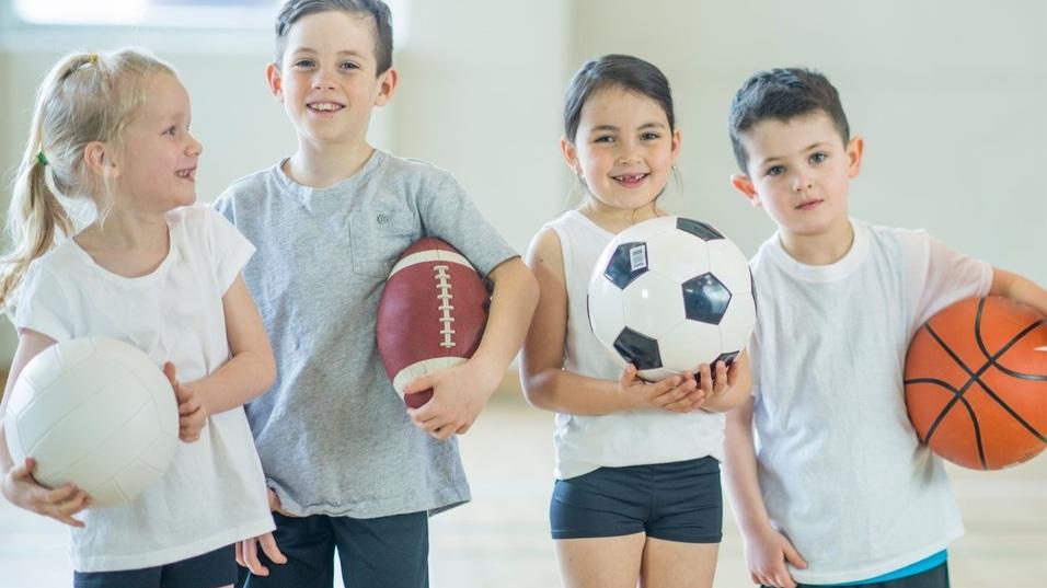 Vier Kinder stehen in einer Sporthalle, halten verschiedene B&auml;lle: Volleyball, American Football, Fu&szlig;ball und Basketball.