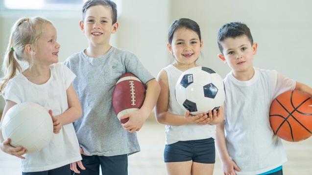 Vier Kinder stehen in einer Sporthalle, halten verschiedene B&auml;lle: Volleyball, American Football, Fu&szlig;ball und Basketball.
