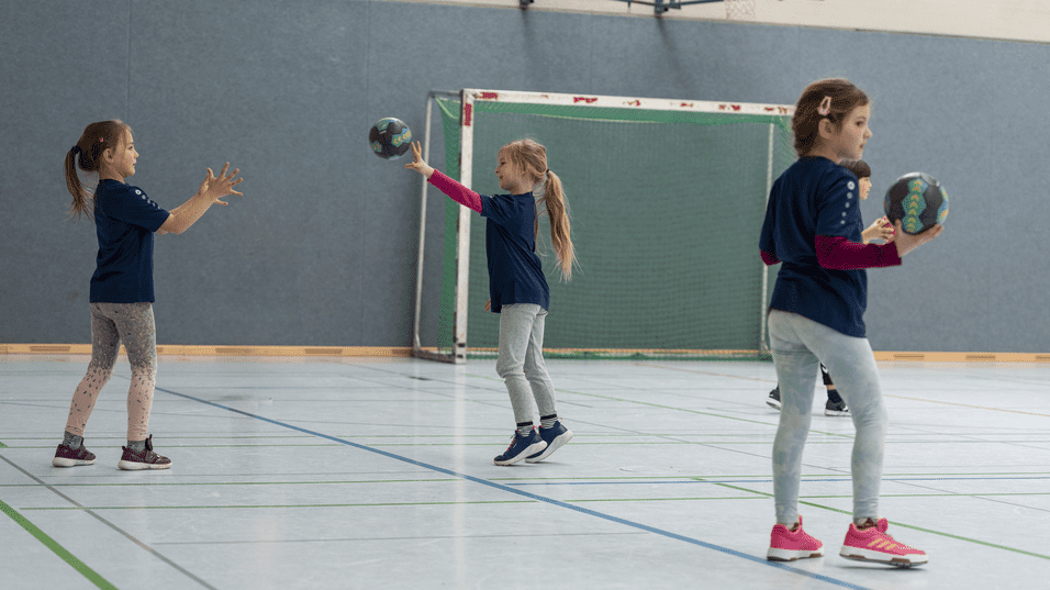 Drei Mädchen in blauen T-Shirts üben das Fangen und Werfen mit Handbällen in einer Sporthalle.
