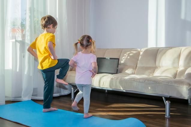 Zwei Kinder stehen barfuß auf einer blauen Yogamatte und schauen auf einen Laptop neben einem Sofa.