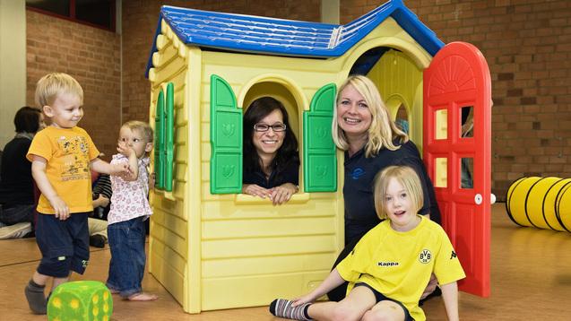 Zwei Frauen und drei Kinder spielen vor einem gelben Spielhaus im Raum mit Holzboden.