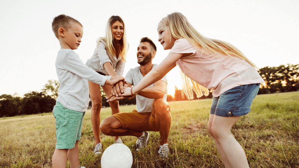 Familie spielt gemeinsam auf einer Wiese, um einen Fu&szlig;ball versammelt, mit H&auml;nden in der Mitte. Sonnenuntergang im Hintergrund.