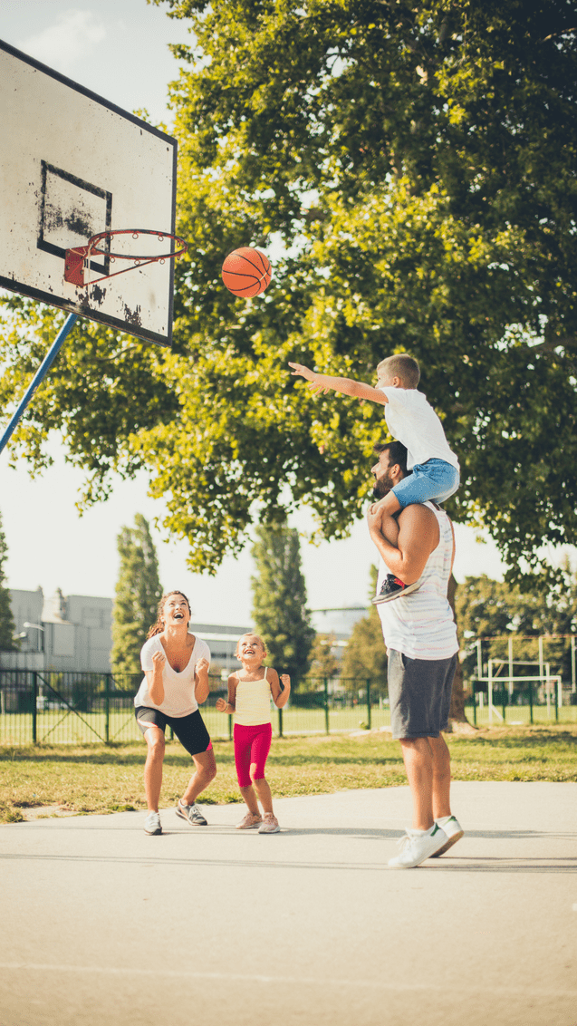 Kind wirft Ball in den Basketballkorb, während er auf einem Erwachsenen sitzt. Zwei weitere Kinder und eine Frau jubeln.