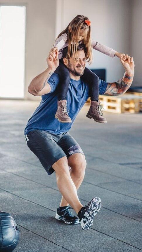 Mann mit kurzer Hose f&uuml;hrt ein Gleichgewichts&uuml;bung mit einem M&auml;dchen auf den Schultern in einem Fitnessstudio aus.