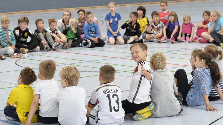 Kinder sitzen in einem Kreis auf dem Boden. Einige sind von vorne, andere von hinten zu sehen. Ein Kind in der Mitte dreht sich zur Kamera um und zeigt mit einem Lächeln den Daumen nach oben. Die Stimmung wirkt fröhlich und entspannt.