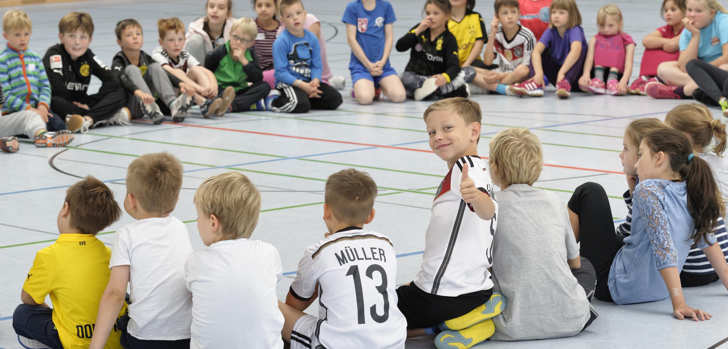 Kinder sitzen in einem Kreis auf dem Boden. Einige sind von vorne, andere von hinten zu sehen. Ein Kind in der Mitte dreht sich zur Kamera um und zeigt mit einem L&auml;cheln den Daumen nach oben. Die Stimmung wirkt fr&ouml;hlich und entspannt.