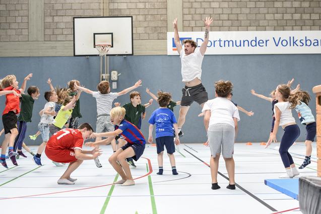 Gruppenspiel mit Kindern in einer Sporthalle, die springen und sich in Bewegung befinden, w&auml;hrend ein Erwachsener anleitet.