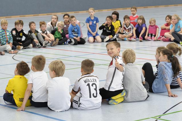 Kinder sitzen in einem Kreis auf dem Boden. Einige sind von vorne, andere von hinten zu sehen. Ein Kind in der Mitte dreht sich zur Kamera um und zeigt mit einem L&auml;cheln den Daumen nach oben. Die Stimmung wirkt fr&ouml;hlich und entspannt.