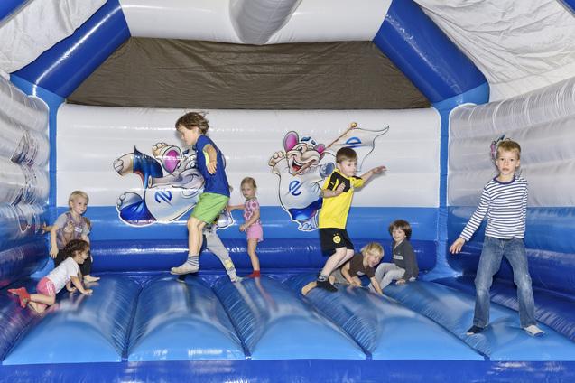 Sechs Kinder springen und spielen auf einer blauen H&uuml;pfburg mit bunten Tiermotiven im Hintergrund.