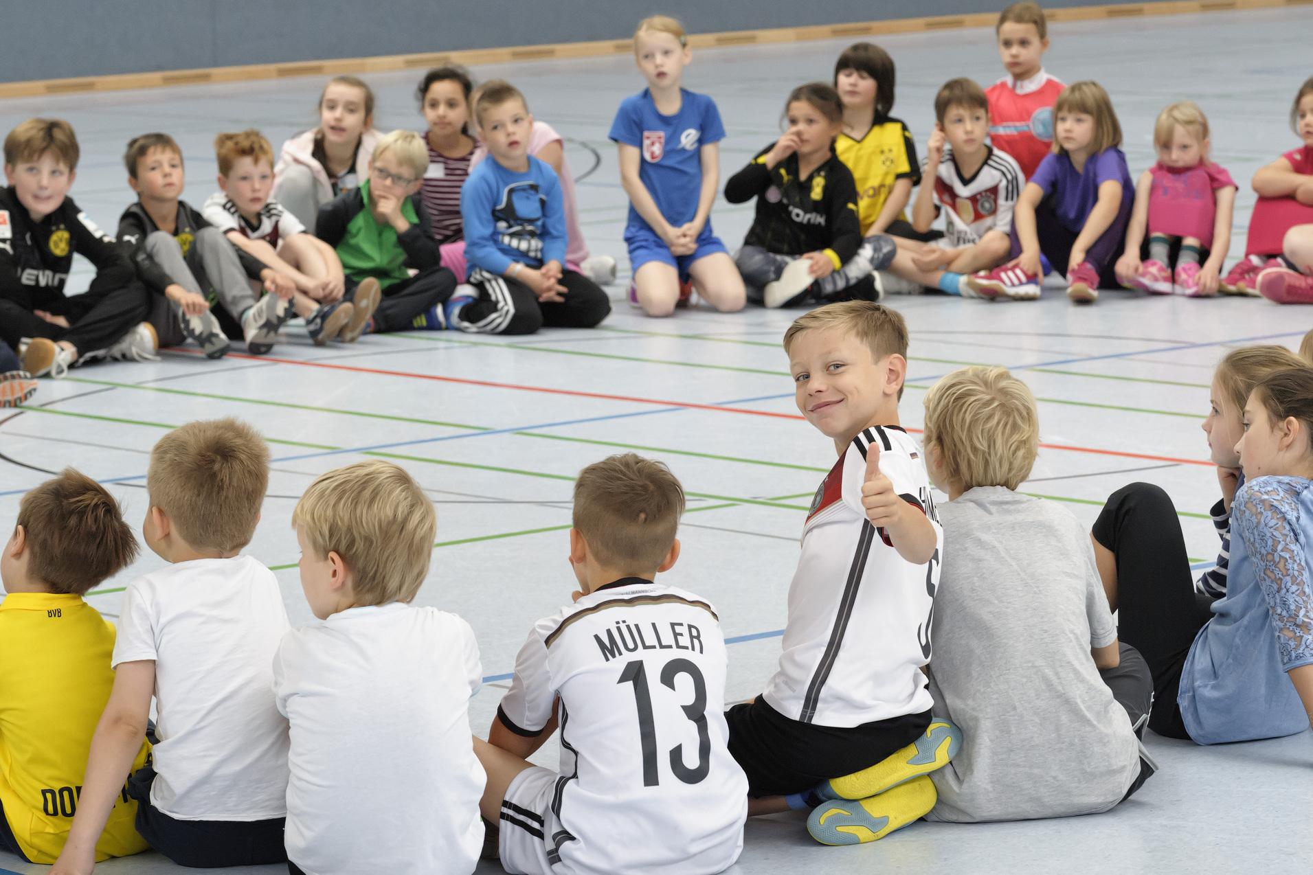 Kinder sitzen in einem Kreis auf dem Boden. Einige sind von vorne, andere von hinten zu sehen. Ein Kind in der Mitte dreht sich zur Kamera um und zeigt mit einem L&auml;cheln den Daumen nach oben. Die Stimmung wirkt fr&ouml;hlich und entspannt.