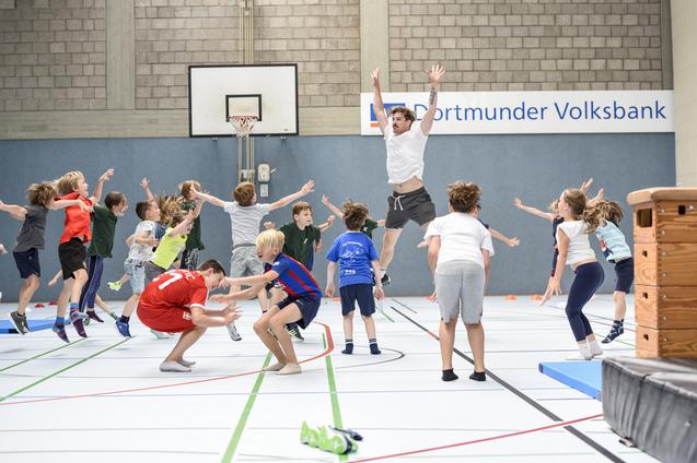 Gruppenspiel mit Kindern in einer Sporthalle, die springen und sich in Bewegung befinden, w&auml;hrend ein Erwachsener anleitet.