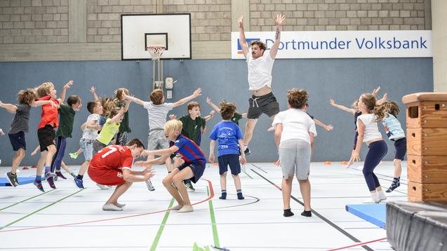 Gruppenspiel mit Kindern in einer Sporthalle, die springen und sich in Bewegung befinden, w&auml;hrend ein Erwachsener anleitet.