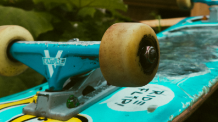 Nahaufnahme eines abgenutzten Skateboards mit blauen Achsen und braunen Rollen, umgeben von gr&uuml;nen Pflanzen.