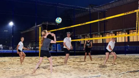 Sechs Personen spielen Volleyball auf einem beleuchteten Sandplatz bei nacht.