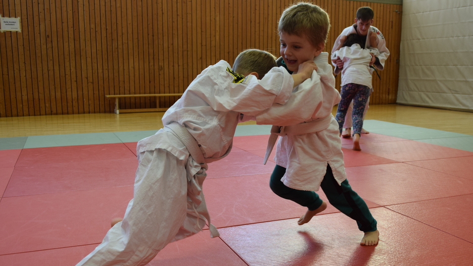 Zwei Kinder im Judogi k&auml;mpfen spielerisch auf einer roten Judomatte, w&auml;hrend ein weiterer K&auml;mpfer im Hintergrund trainiert.