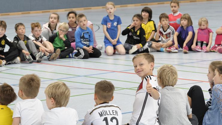 Kinder sitzen in einem Kreis auf dem Boden. Einige sind von vorne, andere von hinten zu sehen. Ein Kind in der Mitte dreht sich zur Kamera um und zeigt mit einem Lächeln den Daumen nach oben. Die Stimmung wirkt fröhlich und entspannt.