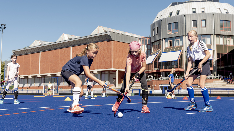 Drei Kinder spielen Hockey auf einem blauen Kunstrasenplatz, im Hintergrund ein modernes Sportgeb&auml;ude.