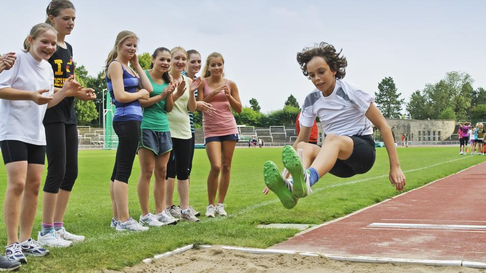 Ein Junge springt im Weitsprung &uuml;ber die Sandschicht, w&auml;hrend eine Gruppe von M&auml;dchen ihn anfeuert.