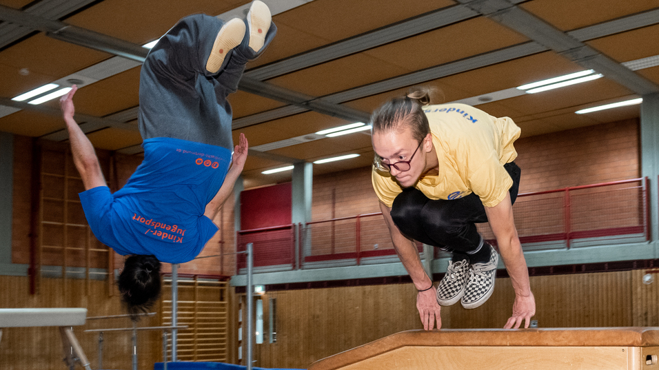 Zwei Turner f&uuml;hren Spr&uuml;nge &uuml;ber ein holzfarbenes Ger&auml;t in einer Turnhalle aus, umgeben von Sportmatten.