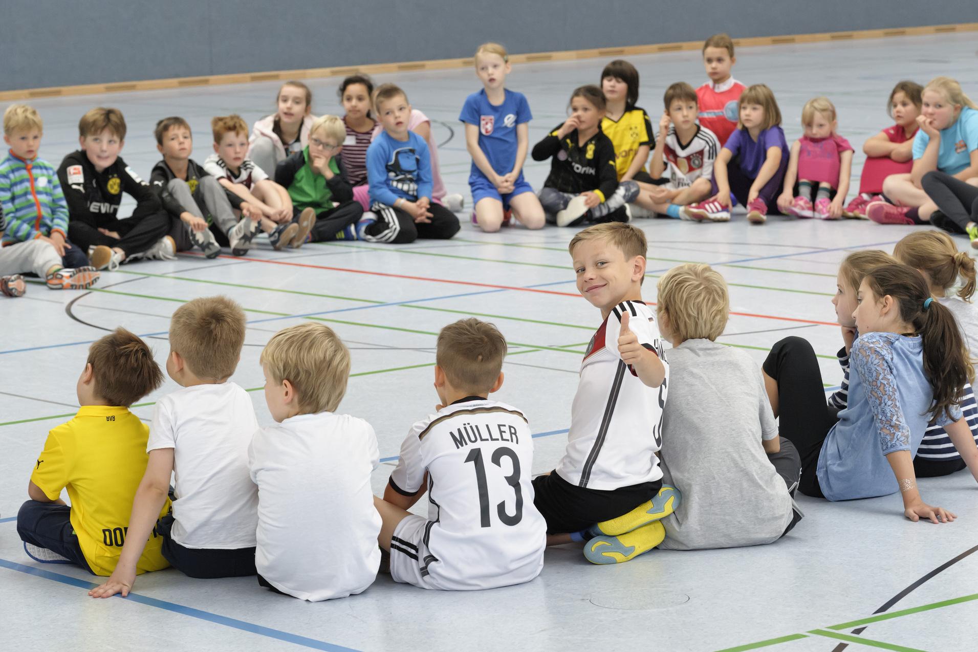 Kinder sitzen in einem Kreis auf dem Boden. Einige sind von vorne, andere von hinten zu sehen. Ein Kind in der Mitte dreht sich zur Kamera um und zeigt mit einem Lächeln den Daumen nach oben. Die Stimmung wirkt fröhlich und entspannt.
