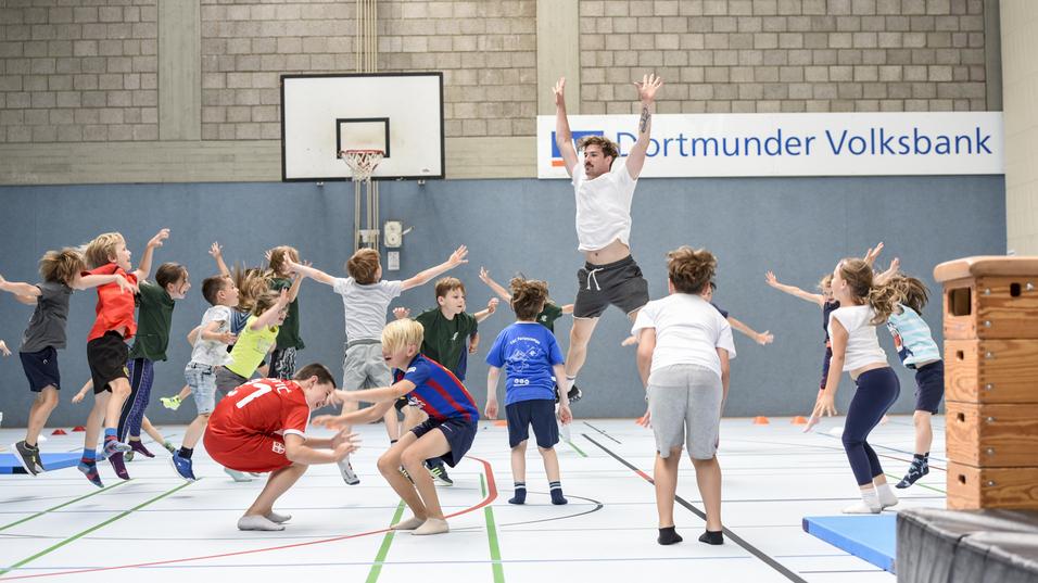 Gruppenspiel mit Kindern in einer Sporthalle, die springen und sich in Bewegung befinden, während ein Erwachsener anleitet.