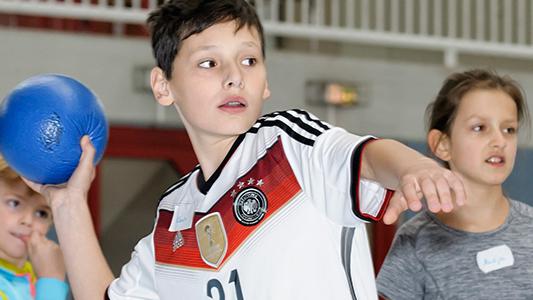 Ein Junge in Deutschland-Trikot wirft einen blauen Ball, umgeben von drei weiteren Kindern in einer Turnhalle.