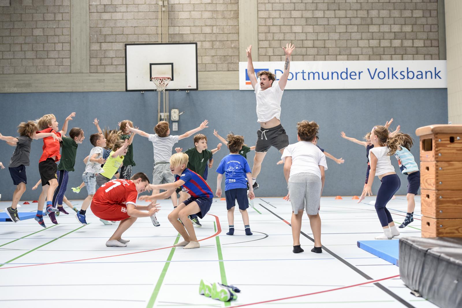 Gruppenspiel mit Kindern in einer Sporthalle, die springen und sich in Bewegung befinden, w&auml;hrend ein Erwachsener anleitet.