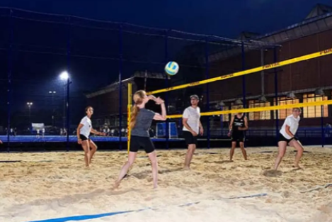 Sechs Personen spielen Volleyball auf einem beleuchteten Sandplatz bei nacht.