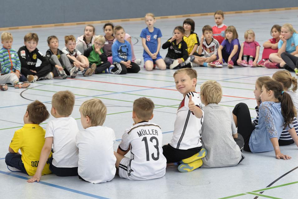Kinder sitzen in einem Kreis auf dem Boden. Einige sind von vorne, andere von hinten zu sehen. Ein Kind in der Mitte dreht sich zur Kamera um und zeigt mit einem Lächeln den Daumen nach oben. Die Stimmung wirkt fröhlich und entspannt.