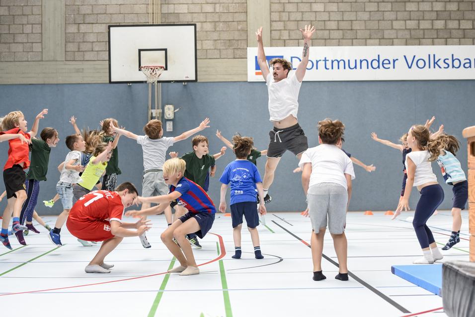Gruppenspiel mit Kindern in einer Sporthalle, die springen und sich in Bewegung befinden, während ein Erwachsener anleitet.
