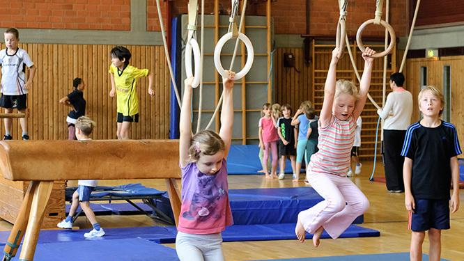 Kinder trainieren in einer Turnhalle an Ger&auml;ten wie Ringen, Barren und auf Matten. Einige spielen und &uuml;ben verschiedene &Uuml;bungen.