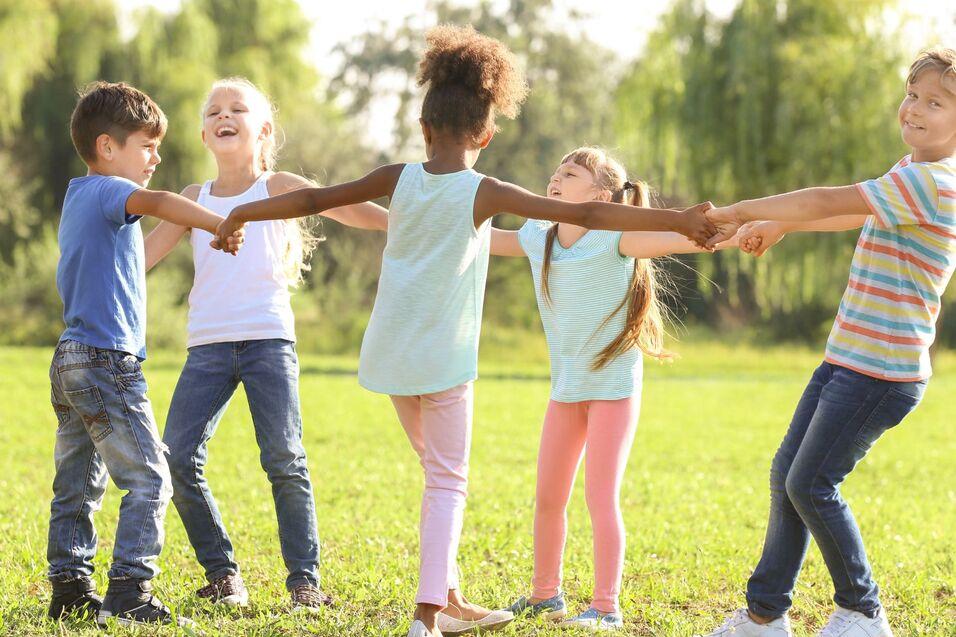 F&uuml;nf Kinder stehen im Kreis auf einer Wiese und halten sich an den H&auml;nden, lachen und spielen zusammen.