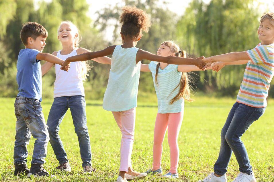 F&uuml;nf Kinder stehen im Kreis auf einer Wiese und halten sich an den H&auml;nden, lachen und spielen zusammen.