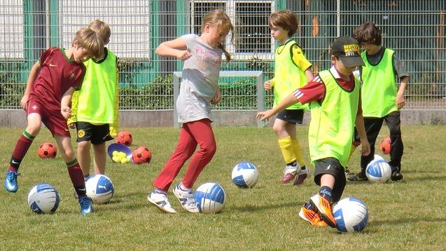 Sechs Kinder in Trainingswesten üben dribbeln mit blauen Fußbällen auf einem grünen Sportplatz.