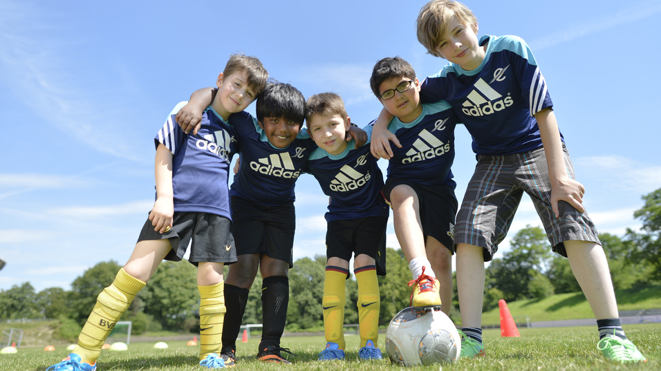 F&uuml;nf Kinder in Sportkleidung stehen lachend zusammen auf einem Fu&szlig;ballfeld, ein Ball liegt vor ihnen.