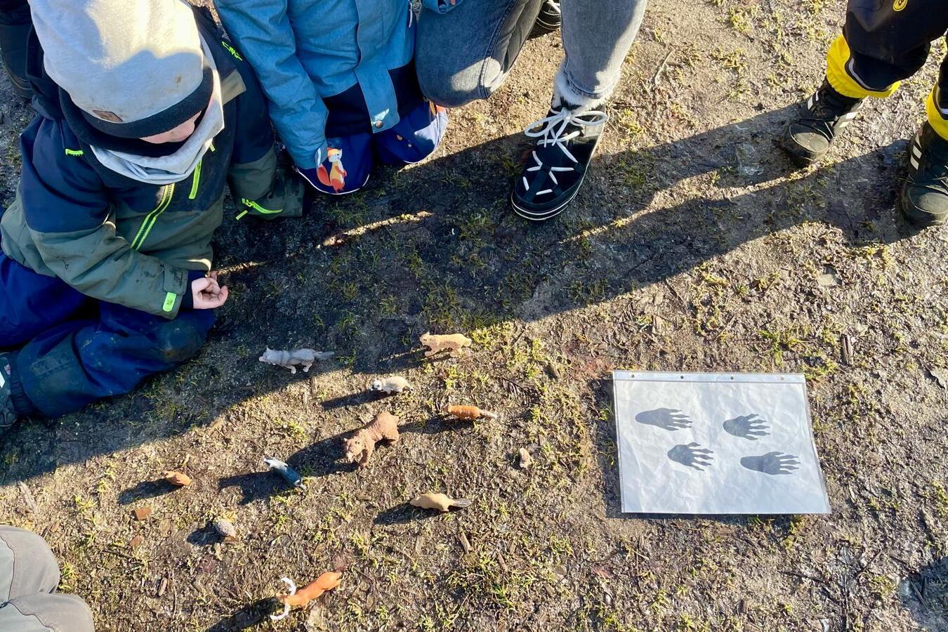 Gruppierte Kinder sitzen um ein Blatt mit Handabdr&uuml;cken, daneben sind bemalte Steine auf dem Boden angeordnet.