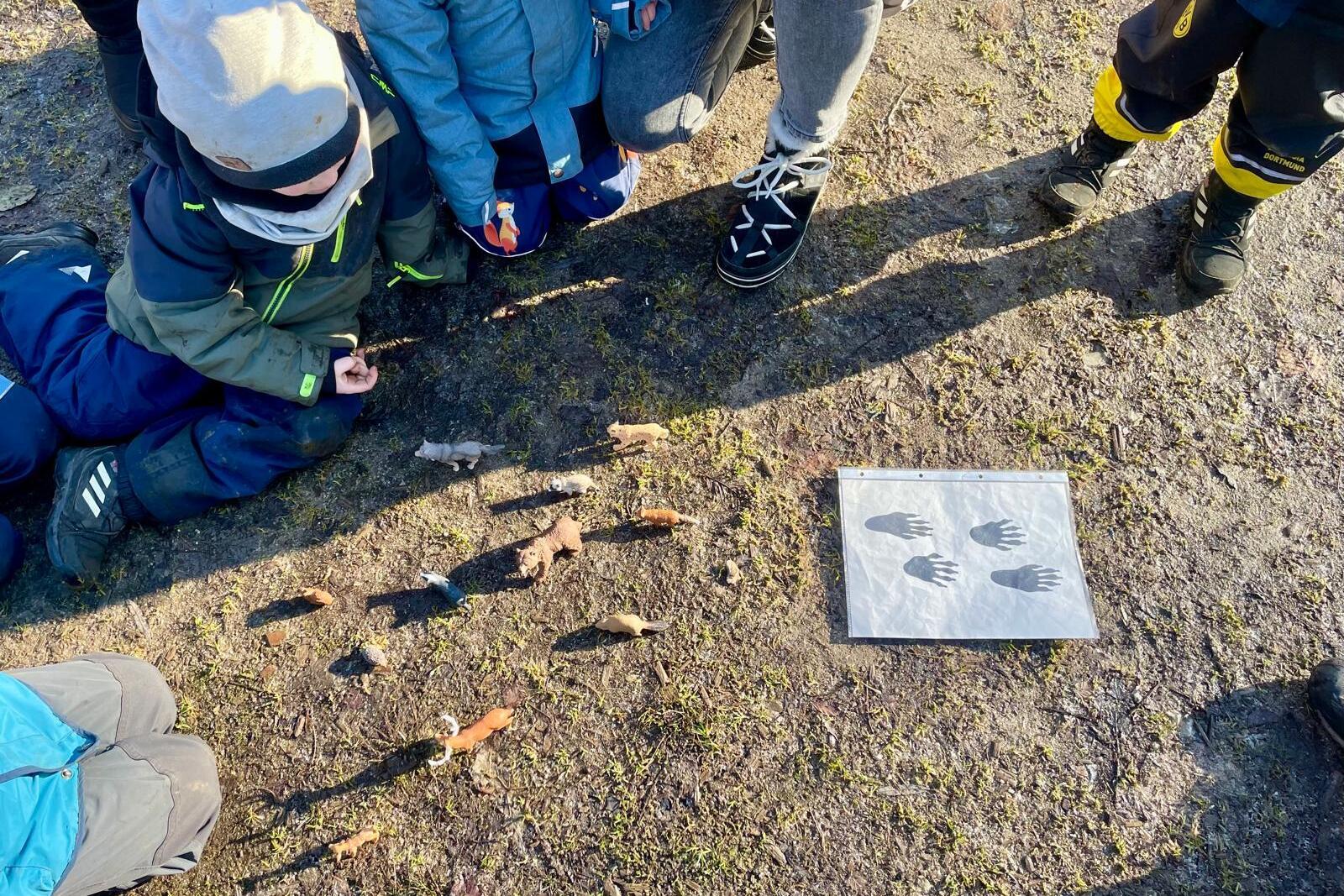 Gruppierte Kinder sitzen um ein Blatt mit Handabdr&uuml;cken, daneben sind bemalte Steine auf dem Boden angeordnet.