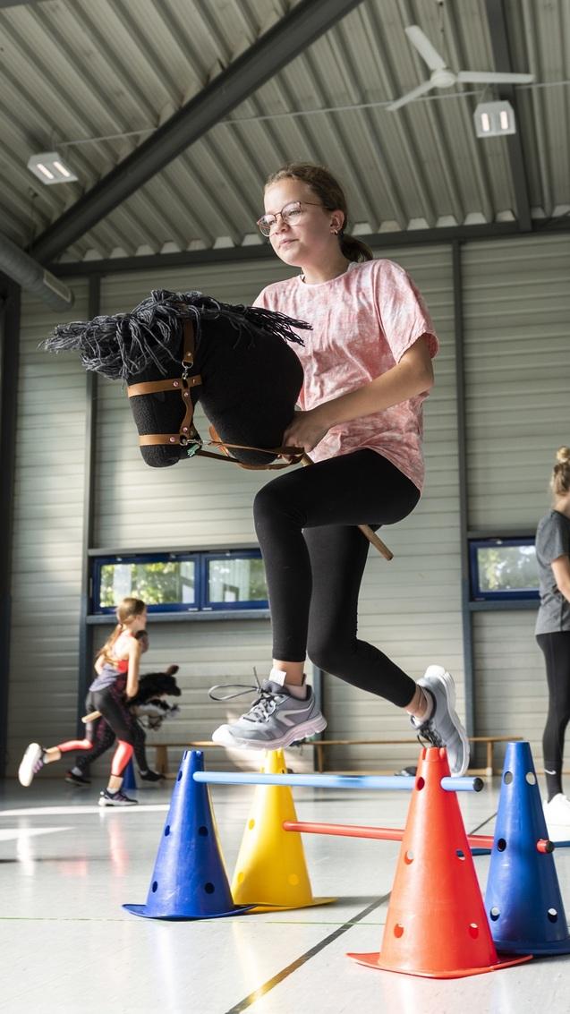 M&auml;dchen springt mit einem Steckenpferd &uuml;ber bunte Kegel in einer Sporthalle, andere Teilnehmer trainieren im Hintergrund.