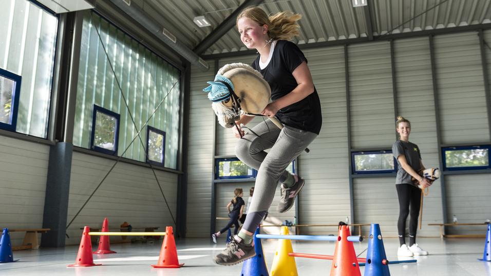 Mädchen springt mit einem Hobby-Pferd über Hindernisse aus Pylonen in einer Sporthalle.
